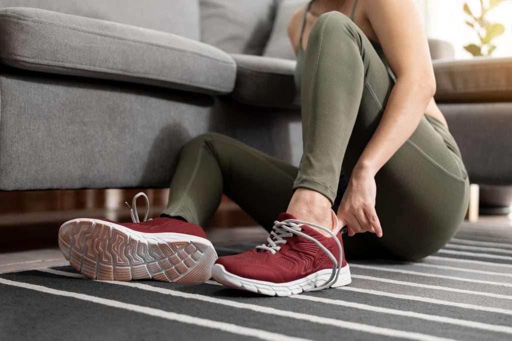 A woman wearing workout clothes puts on her trainers at home, preparing to exercise.